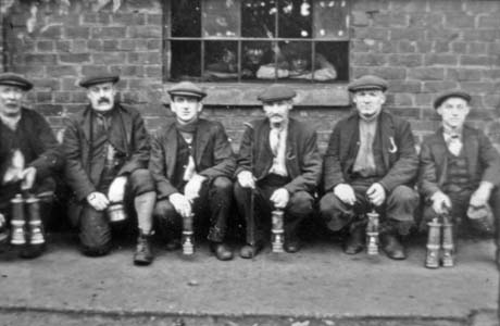 Photograph of six men wearing caps, jackets, scarves, trousers or breeches and boots, squatting against a brick wall in which there is a window through which a man is peering; they are all carrying miner's lamps; they have been identified as being in Shotton