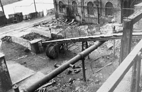 Photograph showing a pipe running across the picture with a chute lying across it and going to the ground; the exterior of a building with round-headed windows is on the right; there is debris on the ground beneath the pipe and chute; beyond the debris is a path, a fence and what seems to be water; the photograph has been identified as being of Shotton Colliery
