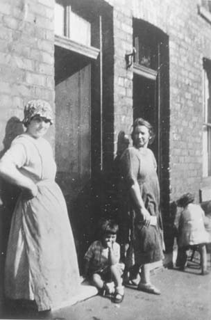 Photograph of a woman wearing a mob cap and a pinafore standing against a brick wall next to a door; a child, aged approximately four years, is sitting on the door step; on the other side of the doorway, is another woman dressed in a pinafore, and the back of a child of a similar age to the one sitting on the doorstep can be seen beyond her; they have been described as Miners' Wives, Shotton