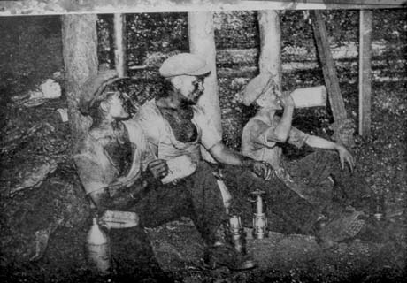 Photograph of three miners underground sitting near pit props; all are wearing caps, shirts, trousers and boots and have pit dirt on their faces and chests; there are two miner's lamps in front of them; the man on the left has a liquid container and a food container near him; the man on the right is drinking from a container; they have been described as miners in Shotton
