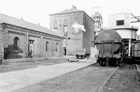 Photograph showing an open space before a colliery building; there are rails running along the space and a coal waggon can be seen on one set of rails and the back of a locomotive on the other; in the middle distance a tall colliery building, presumably the winding house, can be seen with the winding gear just beyond it; the photograph has been described as Colliery Yard, Shotton
