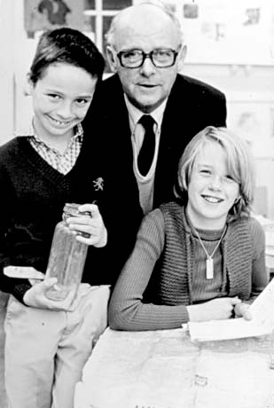 Photograph of a balding middle-aged man wearing glasses, a dark jacket, a dark tie, and a light jumper, standing behind and between boy, aged approximately eleven years, and a girl, aged approximately the same; the boy is wearing long trousers, a dark pullover and a checked shirt; he is holding a jar; the girl is wearing a jumper and a knitted waistcoat and is holding a paper; they have been identified as Mr. Mullen, Headmaster, with Andrew Skinner and Michelle Wheatley