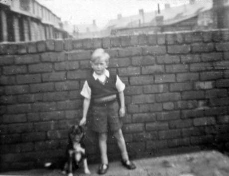 Photograph of a small boy, aged approximately seven years, standing against a wall with a small particoloured dog; the boy is wearing short trousers, short-sleeved shirt, and pullover; roofs can be seen over the wall behind him; he has been identified as being in Shotton