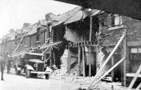 Photograph showing a terrace of houses with two on the middle of the picture with their fronts missing; a motor lorry, registration APT 364, is standing in front of the destroyed buildings with two men in its back and two men standing in the road; a ladder is being put into one of the houses and another ladder can be seen on the roof of a house two doors away; the photograph has been identified as Queen Street After Bombing, Shotton