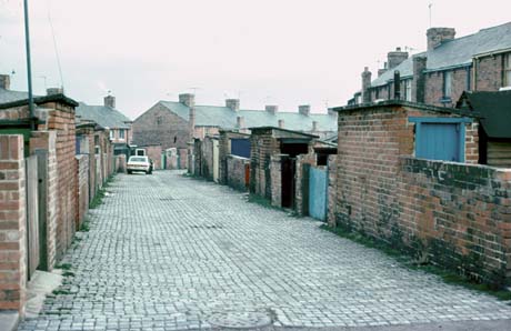 Photograph of the surface of a cobbled street running between the back walls of the yards of terraced houses; a car is parked in the distance in the road; terraced houses can be seen in the distance; the back street has been identified as being in Shotton