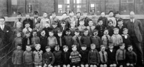 Photograph of forty eight boys, aged approximately seven years, posed in four rows against the wall and windows of a brick building; two men are standing on either side of them; a child in the front row is holding a notice which reads: Shotton Junior School Class 4 1927