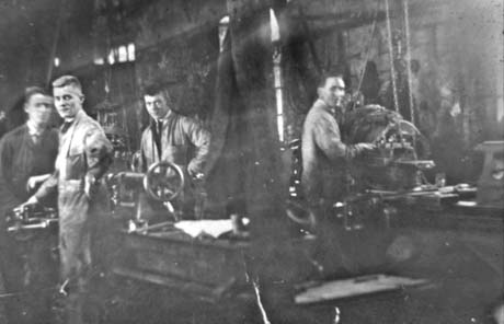 Photograph of four young men in overalls in a building in which the natural light is almost totally excluded; they are standing near benches on which there are machines, one of which has a wheel; they have been identified as Shotton Men At South Hetton Fitter Shop