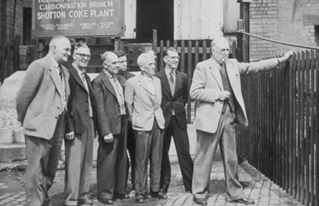 Photograph showing six men standing, watching another man standing with his hand on a gate in a fence, on the right of the picture; behind the men are brick walls, part of the fence and a notice reading: ...Carbonisation Branch Shotton Coke Plant Manager's Office Foreman's Office; the photograph has been described as depicting the closure of the coke ovens at Shotton