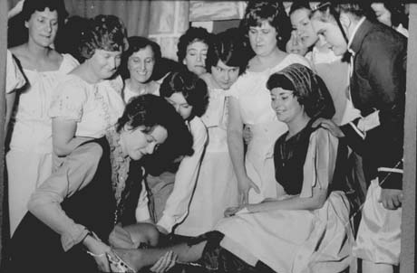 Photograph showing ten women, wearing fancy blouses and plain skirts watching a woman in a jerkin and a ruffled blouse kneeling and putting a shoe on the foot of another woman, who is wearing a skirt, apron and scarf on her head and sitting down; at the side, behind the woman sitting is a woman dressed as a page boy; it is, therefore, likely that the scene depicts characters from a production of Cinderella, which has been identified as taking place in Shotton