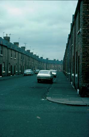 Photograph showing a street of terraced houses curving away to the right; the houses have small gables in the roofs; there are four cars parked in the street, which has been identified as Victoria Street, Shotton