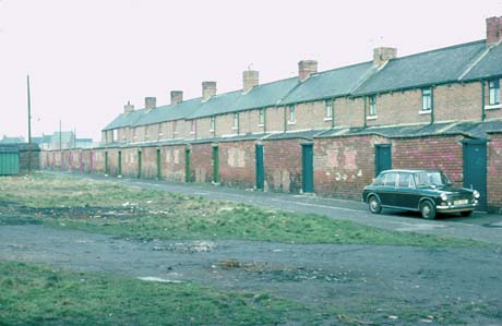 Photograph of the rear of a terrace of houses showing the first floor, roofs of extensions on the ground floor and the walls of the yards of the houses; a car is parked near the back wall and open space with grass and bits of rubbish is in the foreground; the scene has been identified as being in Shotton