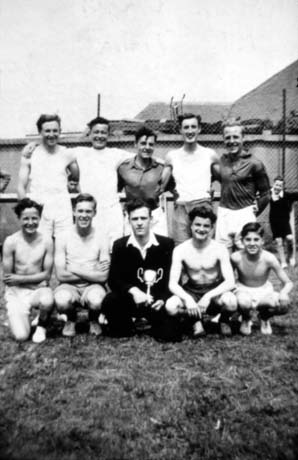 Photograph showing ten young men, posed in two rows in front of a wire fence; nine of the men are in shorts, shirts or bare chests; one is in a blazer and open-necked shirt, holding a trophy cup; they have been described as the Church Lads' Brigade Physical Training Team in Shotton; another young man can be seen in the background watching the team