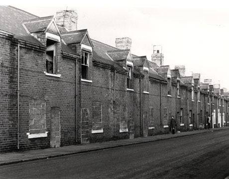 Photograph showing a row of terraced houses with the doors and windows on the ground floor bricked up and the windows on the first floor bricked up or without glass; a woman and a man, carrying a child, can be seen walking along the pavement in front of the houses, which have been identified as being in Shotton