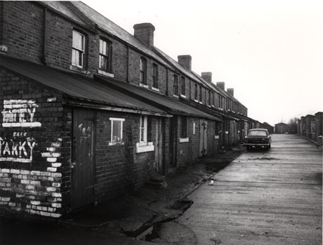 Photograph of the rear of a terrace of brick houses with a lean-to on the ground floor of each house and two windows on the first floor; running the length of the terrace is a road surfaced with rough concrete; a car is parked with its rear to the camera half way along the road; a garage can be seen at the distance and a wall down the right of the picture; the terrace has been identified as being in Shotton