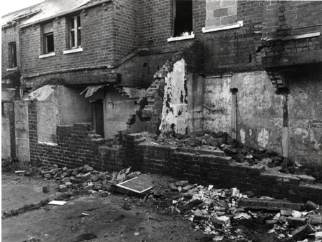 Photograph of houses in the process of being demolished; the facades of two houses, with the ground floor wall partly removed and an internal wall showing, can be seen; two doors and one window can be seen to have been previously bricked up; the houses have been identified as being in Shotton