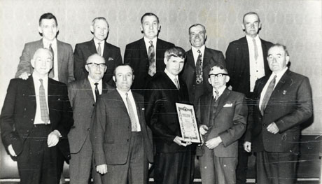 Photograph of eleven men in suits posed in two rows against a wall; a man on the front row is holding a certificate; the photograph has been identified as South Hetton Miners' Presentation, 1971