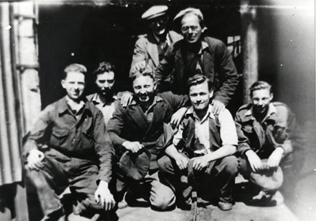 Photograph of five men in overalls and work clothes squatting on the ground with two men standing behind them; they are described as being miners at South Hetton Colliery in 1958