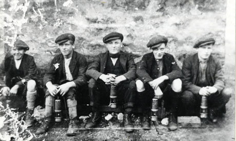 Photograph of five young men sitting in a row with their backs to a rough stone wall; all five are wearing caps, jackets, waistcoats, boots and long socks and are carrying miners' lamps; they have been identified as miners at South Hetton Colliery and, individually, as follows: at the right: J. Pearson; centre: W. Pickering