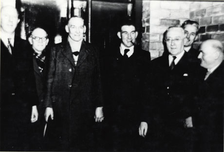 Photograph of seven men wearing overcoats standing in front of a brick wall and a doorway; they have been identified as marking the opening of the pithead baths at South Hetton Colliery in 1954; the following individuals have been identified: Right: Sam Watson; Second Right: Mr. Mckillhope; Third Left: Mr. Bennett; Second Left: Mr. F. Hutchinson