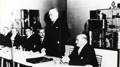 Photograph of five men sitting at a table listening to a sixth man who is standing up making a speech; behind the men is the indistinct figure of another man in a doorway on the left of the picture and a counter with an urn on it; the speaker has been identified as Sam Watson opening the pithead baths, canteen and medical centre at South Hetton Colliery in 1954