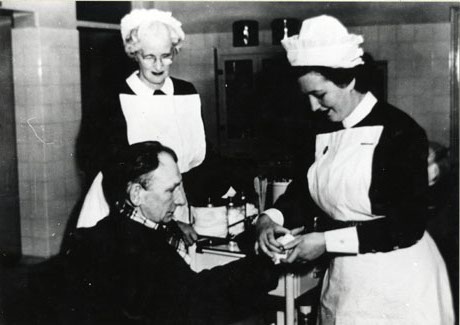 Photograph of a nurse standing at the right of the picture bandaging the hand of a man who is sitting facing her; behind the nurse is a trolley holding medical supplies, and a second nurse is behind that, watching the treatment being given by the first nurse; the photograph has been identified as showing Sister Yeomans in the medical centre at South Hetton Colliery in 1954