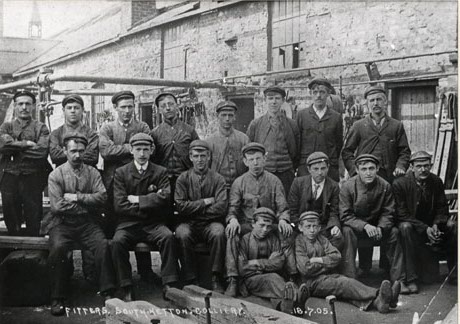 Photograph entitled Fitters, South Hetton Colliery. 18. 07. 05, showing nine men standing, seven men sitting on a bench and two men sitting on the ground with a building of rough stone behind them; the men are all dressed in working clothes except for one man who is dressed in suit, collar and tie; the building behind the men has machinery and equipment attached to it, and is, most likely, a colliery building
