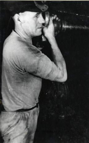 Photograph of a man standing sideways to the camera dressed in miner's helmet, short-sleeved vest and trousers and holding a large screw-like drill at eye level; the photograph has been identified as Using a drill at South Hetton Colliery, 1950