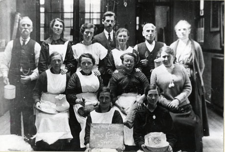 Photograph of eight women wearing aprons, two women wearing skirts and cardigans, two men in shirt sleeves and one man in a suit, grouped inside what may be a school hall; the women in aprons and the men in shirt sleeves are holding bowls, jugs, bread and mugs; a woman on the front row is holding a notice reading: South Hetton Council Girls Canteen Staff; the date given to the photograph is 1926, so it is possible that the canteen staff were providing relief during the strike of that year