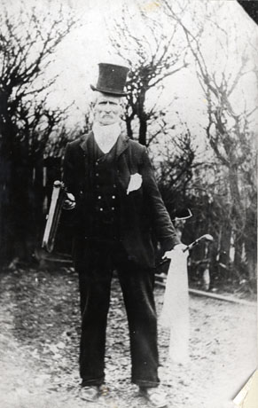 Photograph of an elderly man wearing a top hat, a suit with double-breasted waistcoat and a scarf round his neck, with trees behind him; he is holding a large rattle in his right hand and a stick and cloth in his left; he has been identified as Joe Robson, South Hetton, Crakeman