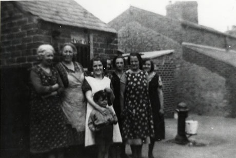 Photograph of seven women and a small boy standing in front of a small outhouse with the side of a low house in the background; behind the group, also, is a stand pipe and bucket; the women are all wearing overalls or aprons; they have been identified as being in the Eight Rows at South Hetton