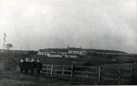 Photograph showing a railway line on the left of the picture and a group of low buildings in the centre set in fields; in the foreground is a fence running across the picture with four boys, aged approximately seven to ten years and wearing suits and Eton collars, standing next to it; they have been identified as Joe, Tom amd William Pine before Patterson's Farm, South Hetton