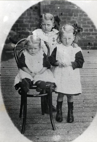 Photograph of a small girl, aged approximately five years wearing boots a dark dress and a broderie anglaise pinafore, is standing beside a wooden dining chair, on which a small girl, aged approximately three years and wearing the same costume as her companion, is sitting; behind both children a third small girl, aged approximately eight years and wearing similar clothes, is standing; behind all three is a brick wall; they have been identified as The Liddle Children in Smith Street, South Hetton, circa 1920