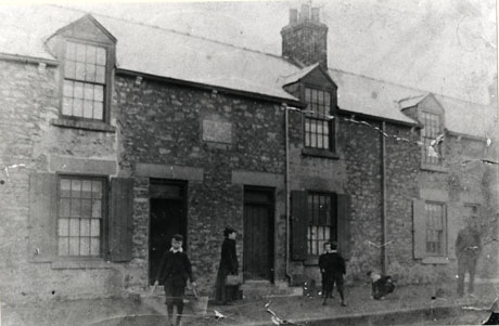 Photograph of the facade of a terrace of houses built of rough stone, one of which has a plaque set into its wall; the building has been identified as The Nurses House South Hetton; in front of the building are a woman, a man and four boys, one of whom, identified as R. Henderson, is carrying a water bucket using a framework described as a girth