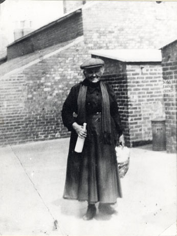 Photograph of a woman dressed in a blouse, a long skirt, a man's cap, and a scarf, standing in a yard with the brick walls of houses behind her; she is carrying a bottle of milk and a basket and has been identified as Mrs. Cook standing in The Eight Rows, South Hetton