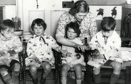 Photograph showing two small boys and two small girls aged approximately five years sitting in small chairs facing the camera; the two boys and one of the girls are wearing patterned overalls; the fourth child is holding a small doll; a young woman, also wearing a patterned overall, is leaning over the small girl with the doll and tipping something from a container into the hands of the small boy beside her; behind the group, a door, a bowl of hyacinths and pictures of two dancing birds or animals can be seen; the photograph has been identified as The Hall, South Hetton, 1938, in use as a Training Centre