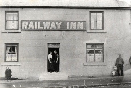 Photograph of the exterior of a building rendered in concrete and showing four windows, a door and a sign reading Railway Inn; faces can be seen at the windows in the ground floor; a man and a woman are standing in the doorway, above which is written: Edward Palmer, Licensed To Retail Ales Wines Spirits and Tobacco; a man with a dog, and a child, are standing on the pavement in front of the building