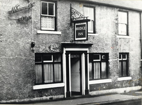 Photograph of the exterior of a building covered in stucco with three windows on the ground floor and three on the first floor; above the doorway is a hanging sign reading Bridge Inn; an electric sign reading Nimmo's Ales is on the wall; the building has been described as The Old Bridge Inn, South Hetton