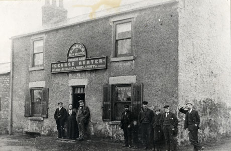 Photograph of the exterior of a building covered in stucco with two windows in the ground, and two windows on the first floor; above the doorway is a sign reading S. H. R. C. C. Head Quarters Butchers Arms George Hunter Licensed to Retail Ales, Wines, Spirits and Tobacco; a woman and two men are standing in the doorway and four men and two boys are standing at the corner nearest to the camera; the public house has ben identified as being at South Hetton