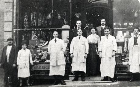 Photograph of four men in white coats and aprons, one boy in a white coat and apron, two men in suits, a woman in a blouse and long skirt, and a man in an informal coat and cap, standing outside the large windows of a shop selling high quality groceries; the contents of the windows can be seen; the name Wakefield is engraved below the window; the shop has ben identified as being in South Hetton
