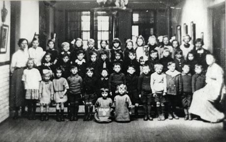 Photograph of forty children aged approximately eight years posed in a room with large windows behind them and bare floorboards below them; they are accompanied by two women who have been identified as: Left: Miss Smithson; Right: Miss Wiseman; a child at the front is holding a notice which can be read only as Group; the children have been identified as attending South Hetton Council School