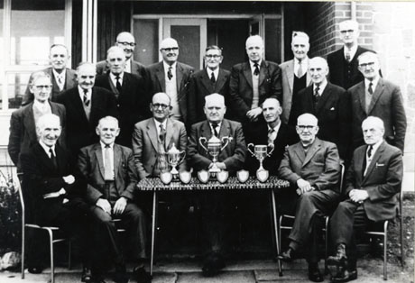 Photograph of nineteen elderly men standing and sitting round a table on which there are three trophy cups and six small shields; all the men are wearing suits and ties; the table and the men are outside the entrance to a building; they have been identified as taking part in a presentation of bowls trophies at South Hetton