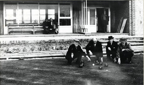 Photograph of four elderly men squatting on a bowling green near its edge, with eight bowling balls and a jack in front of them; behind them are two men sitting on a seat in front of a picture window of a building on the edge of the green; the doorway of the building can also be seen; the green and building have been identified as Welfare Park, New Pavilion and Bowling Green, South Hetton