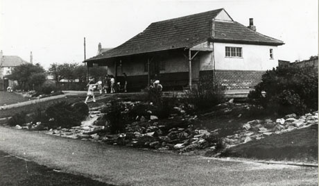 Photograph of a single-storey building with a veranda showing the side and front; indistinct children are playing in front of the building on the veranda and on what appears to be a rockery; a path is running across the front of the of the picture; the building has been identified as the Old Pavilion, Welfare Park, South Hetton