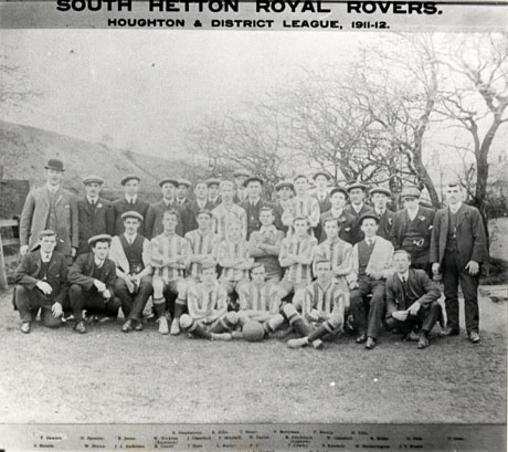 Photograph of eleven men in football strip accompanied by twenty men in suits and posed in the open air in front of trees and what may be a railway embankment; the picture is captioned as follows: South Hetton Royal Rovers Houghton and District League, 1911-12; the individuals in the photograph are named as follows: Back Row, left to right: R. Stephenson; R. Ellis; T. Horner?; F. Berryman; F. Burnip; H. Ellis; Middle Row: F.[ ]; W. Spooner; R. Jones; W. Wickins ? ( Secretary); J. Glassford; F. Mitchell; W. Taylor; R. Ditchburn ( Captain); W. Glassford; R [ ]; G. [ ]; F. [ ]; Front Row: S. Shields; W. Brown; J. A. Anderson; R. Cowey; J. Stott; L. Rutter; F. F.(sic); T. Cowley; S. Kennedy; W. Hetherington; J. T. [ ]