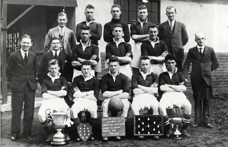 Photograph of eleven men in football strip posed, with five other men in suits, against the wall of a building; in front of the group are two trophy cups and a shield and a tray on each of which twelve medals are pinned; also at the front is a notice which reads: South Hetton Coll. Res. Winners of The Sunderland J. D. C. Amateur League and Hetton Charity Cup, 1931-32