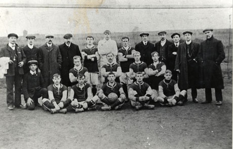 Photograph of twelve men in football strip posed, with ten other men in overcoats, in front of a net or wire fence, with fields behind it; they have been identified as Royal Rovers Football Team, South Hetton, although the strip worn by the players is different from that to be seen in the photograph of the Royal Rovers in 1911 also in the collection