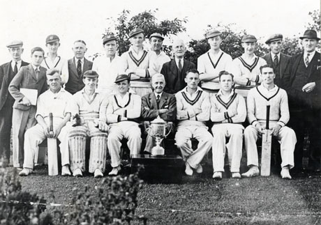Photograph of eleven men in cricket whites posed, with seven men in suits and a boy, in the open air; in front of the group is a trophy cup; the group has been identified as C. W. Cricket Club, Winners of Durham Aged Miners' Cup (Matthew Oswald Cup)