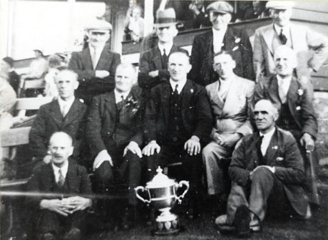 Photograph of eleven men in suits sitting near benches on which spectators can be seen sitting; behind the men is a small wooden building out of the window of which people can be seen looking; in front of the men is a trophy cup; they have been described as Cricket Club Men's Committee at Castle Eden; the cricket club has been identified as that of South Hetton
