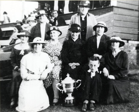 Photograph of nine women sitting near benches on which spectators can be seen sitting; behind them is a small wooden building from the window if which people can be seen looking and in front of them is a trophy cup; the group has been identified as Cricket Club Women's Committee at Castle Eden; the cricket club has been identified as that at South Hetton