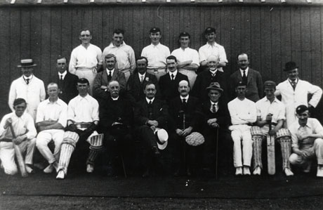 Photograph of eleven men in cricket whites accompanied by ten men in suits and an umpire in his white coat; the group is photographed against the wall of a low wooden building; the group has been identified as the cricket team at South Hetton